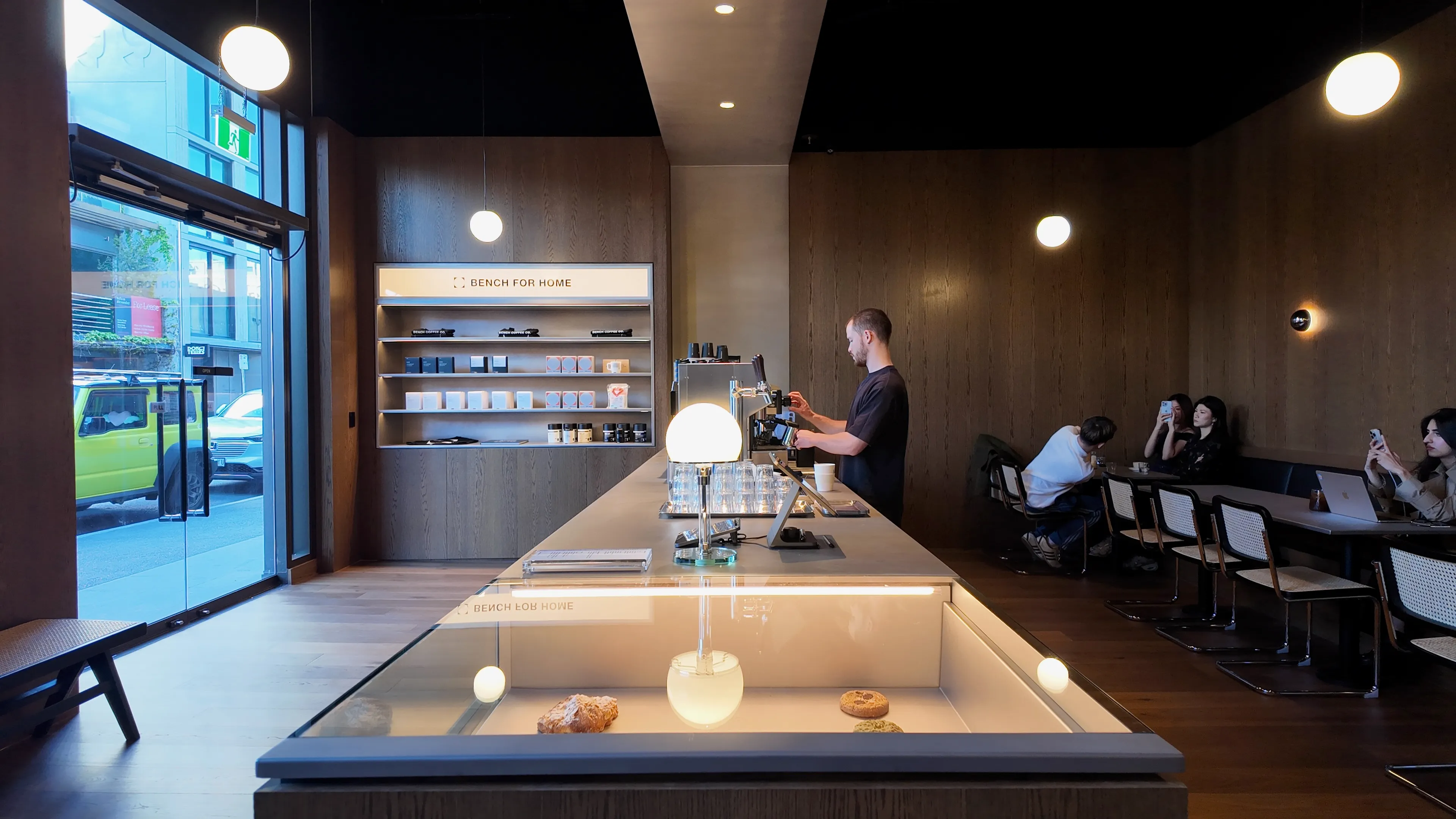 Inside a café with a long counter, a barista at the machine and people at tables.
