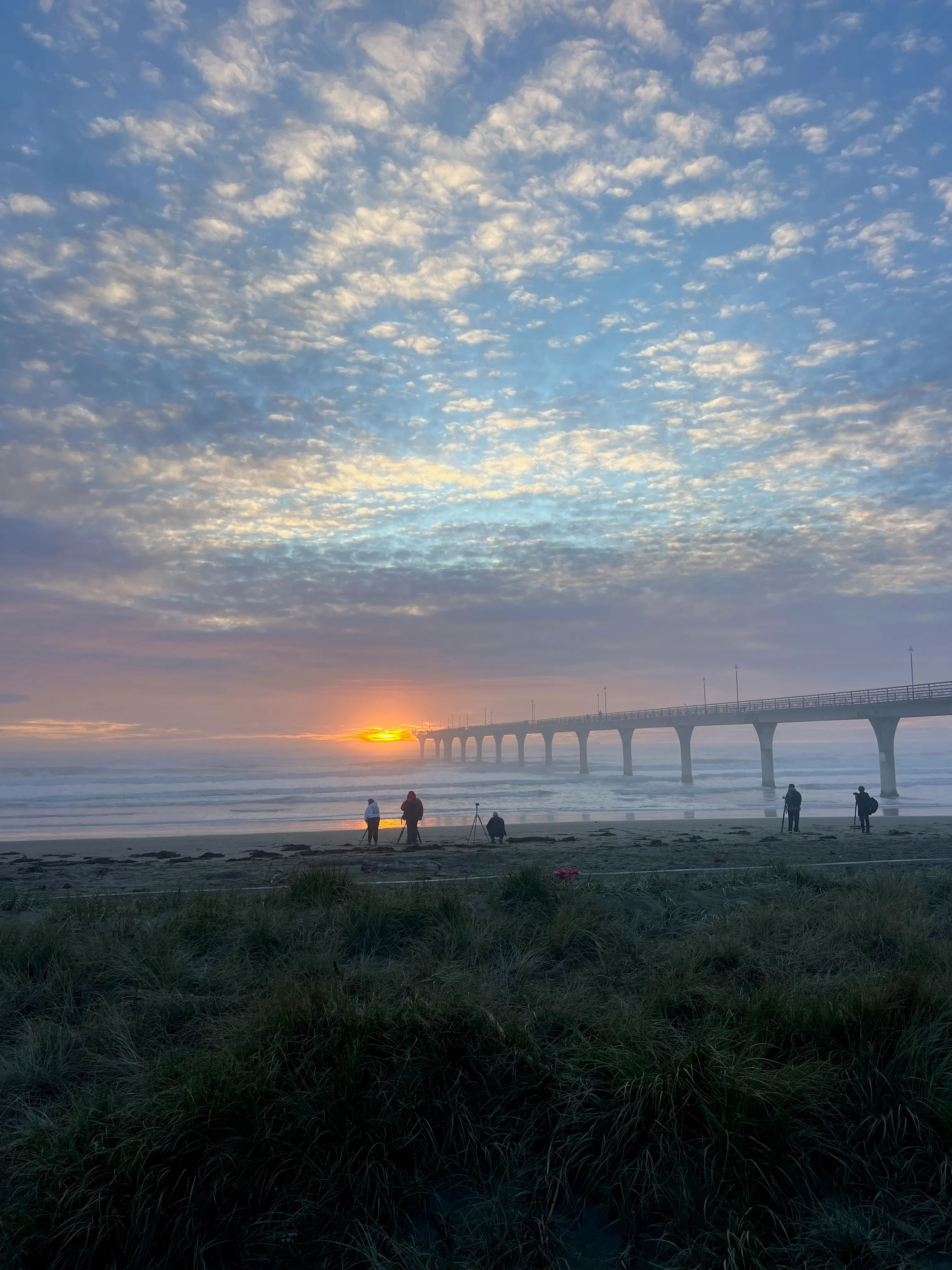Walkers on the sand below a long pier as the sun rises over the sea.