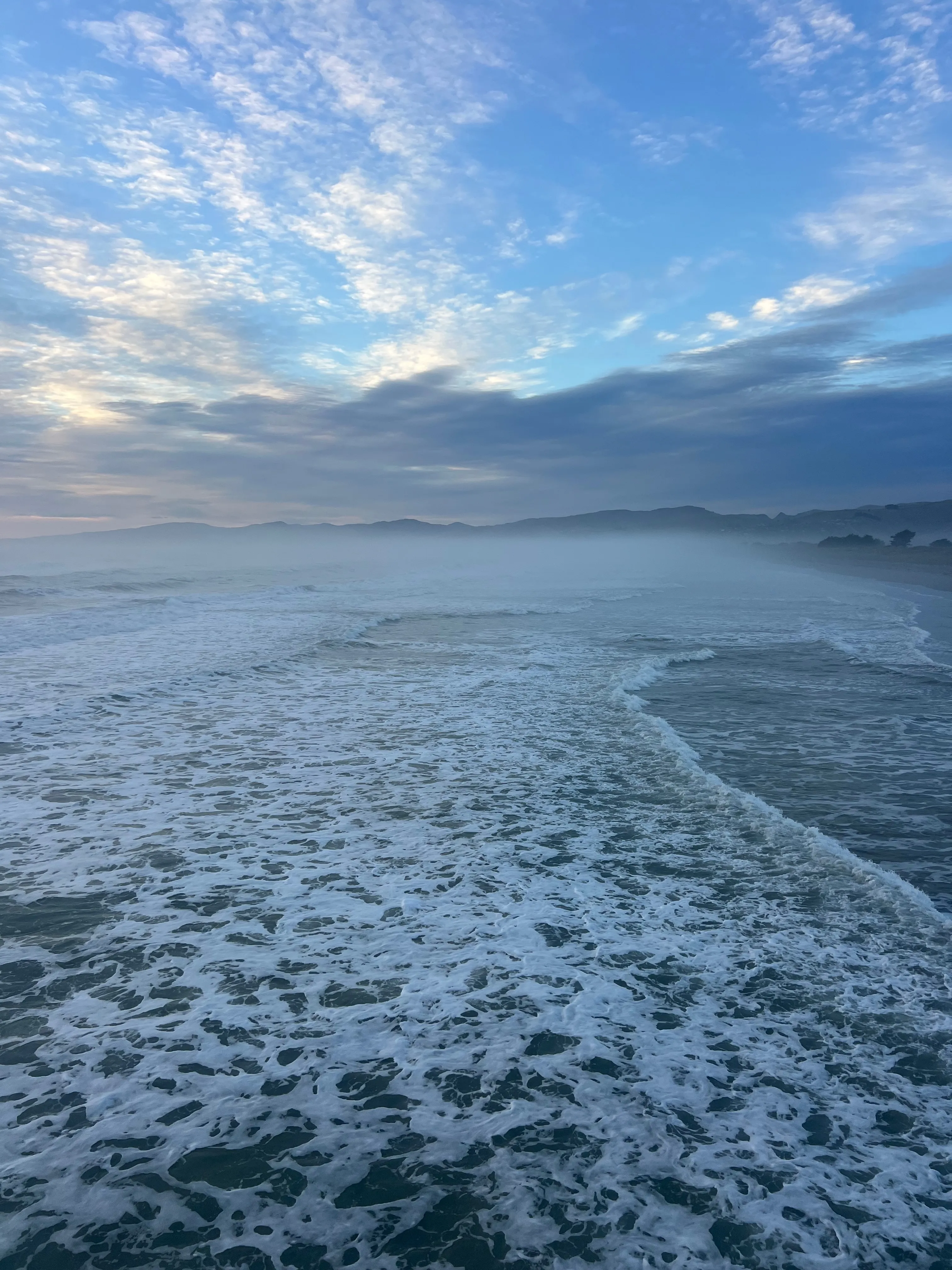 Waves washing onto a misty beach under a streaky blue sky.