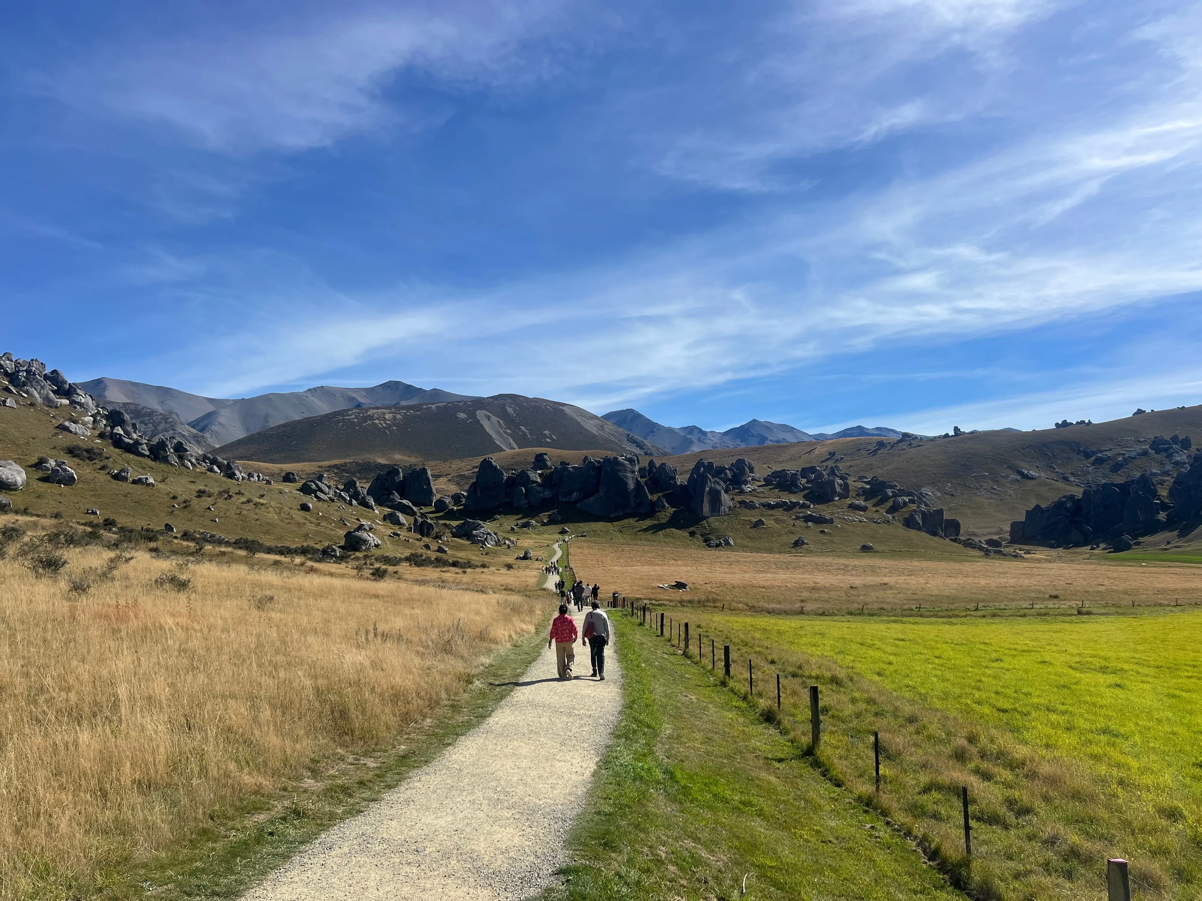 Two people walking a gravel path through golden grass towards limestone boulders and mountains.