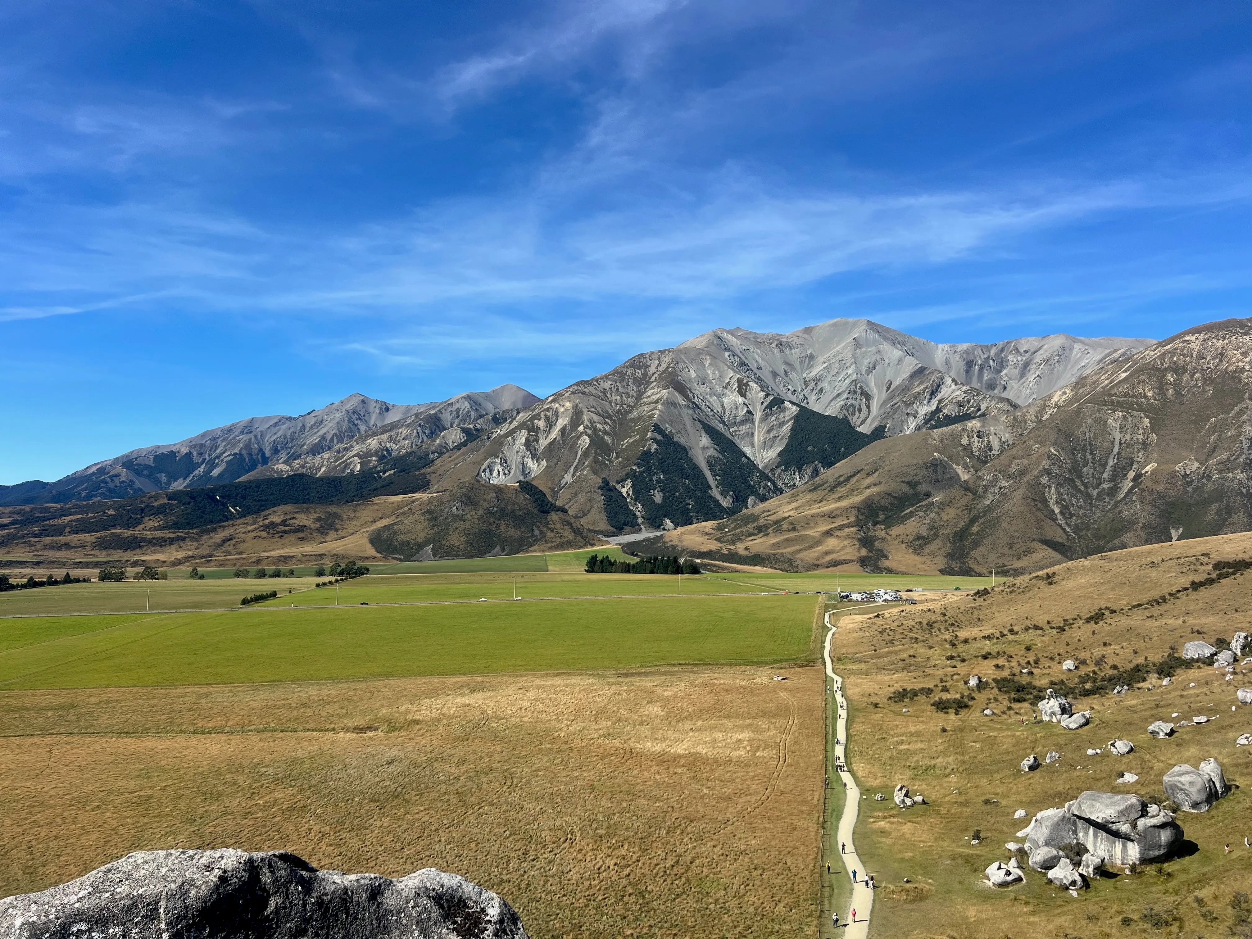 A wide valley of green and golden fields beneath a mountain range and blue sky.