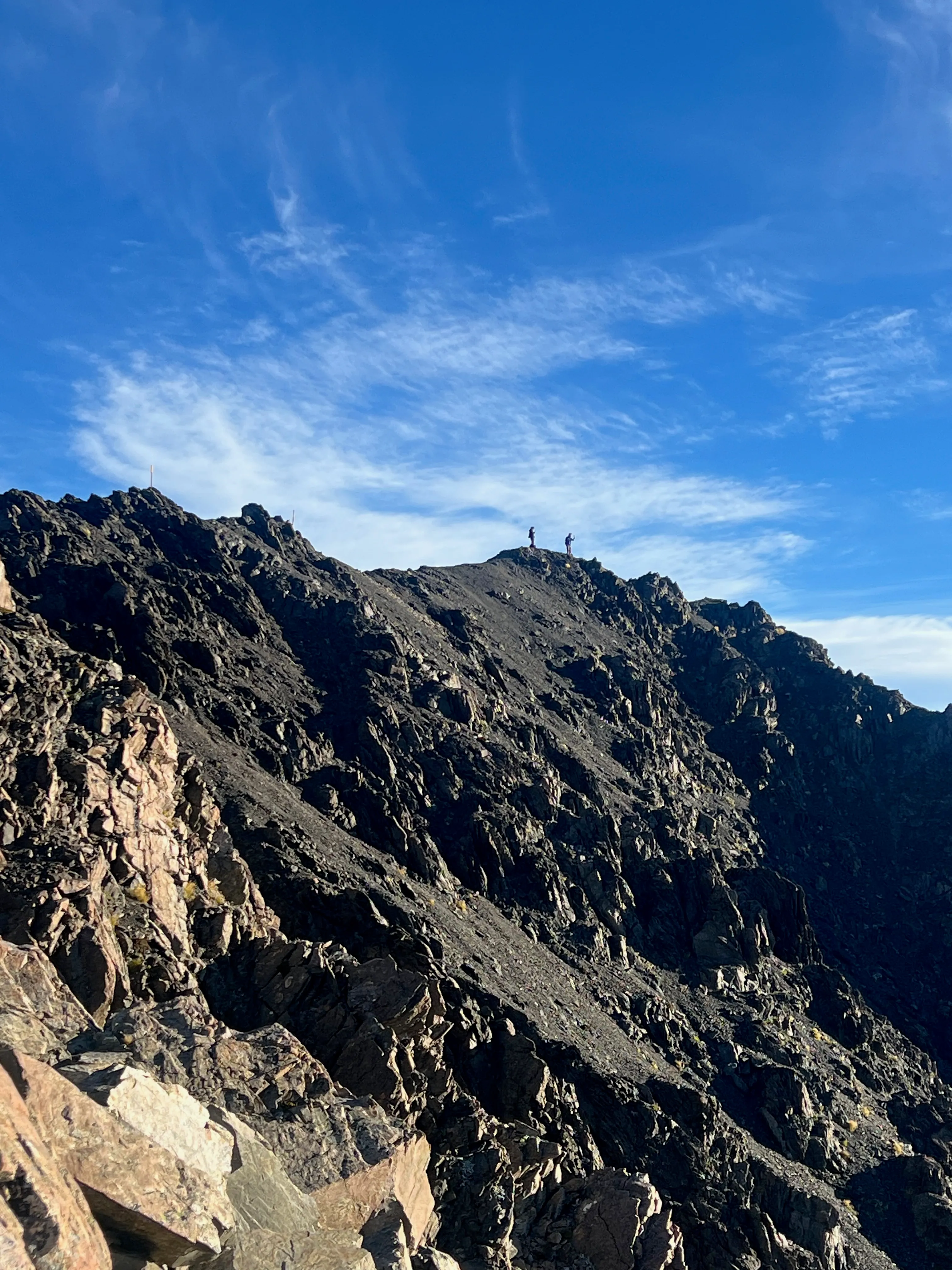 Two small figures standing on top of a dark rocky summit ridge.