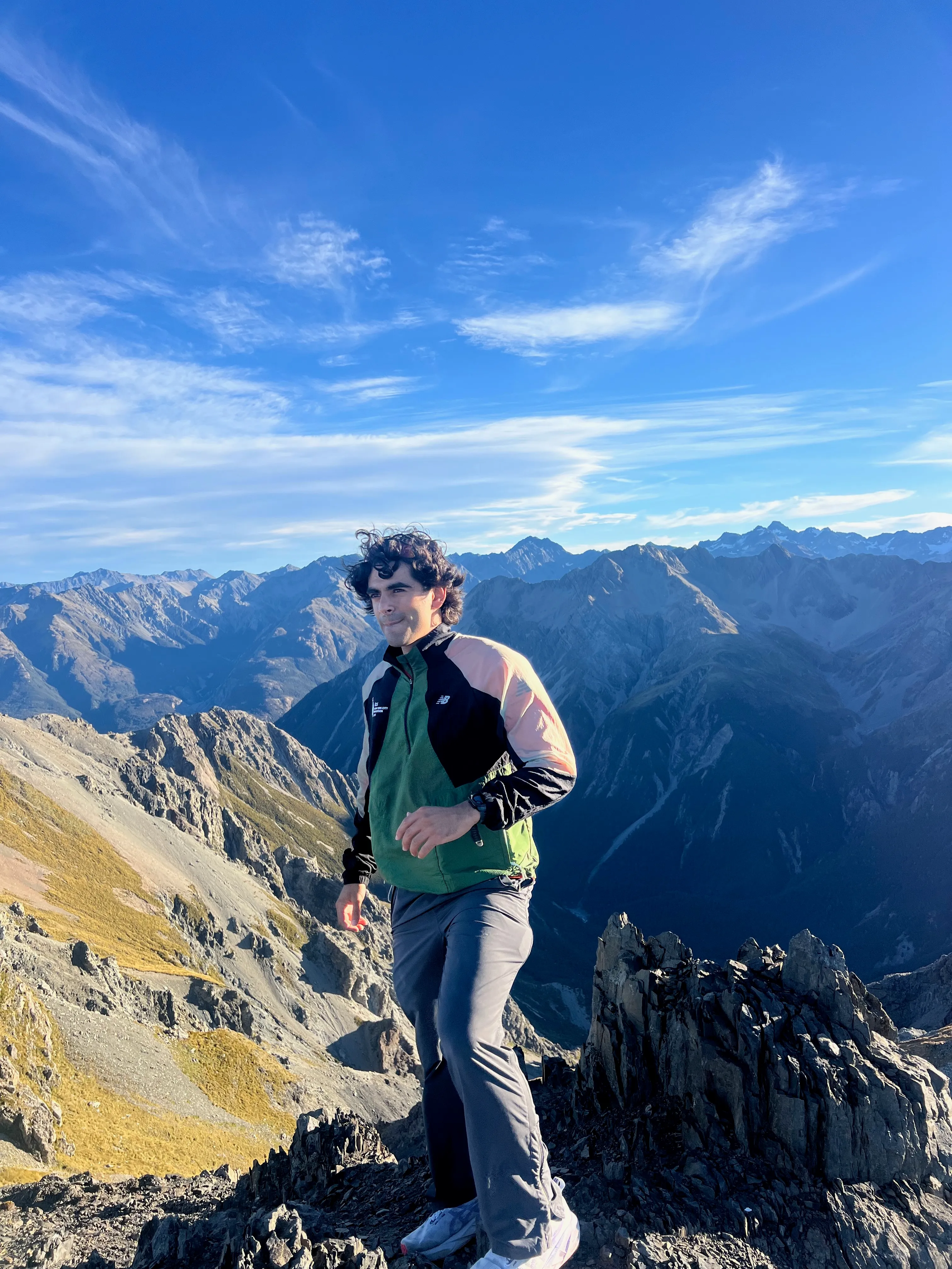 A hiker in a green jacket standing on a rocky summit above mountain ranges.