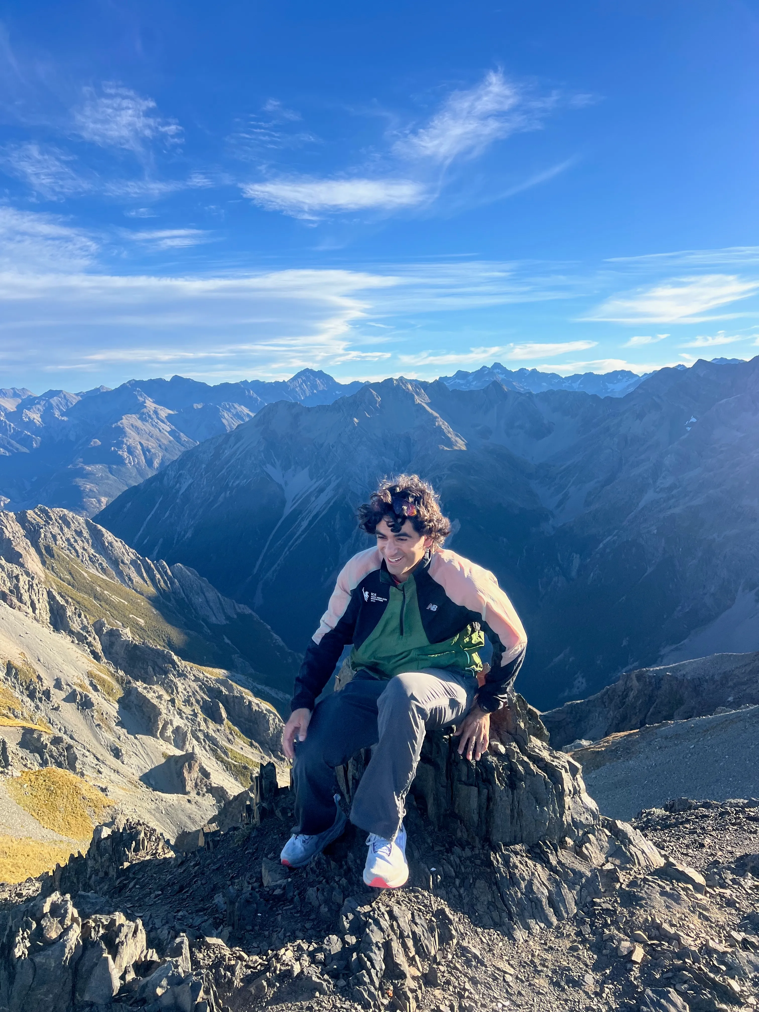 A hiker sitting on a rock on the summit, smiling, with peaks behind.
