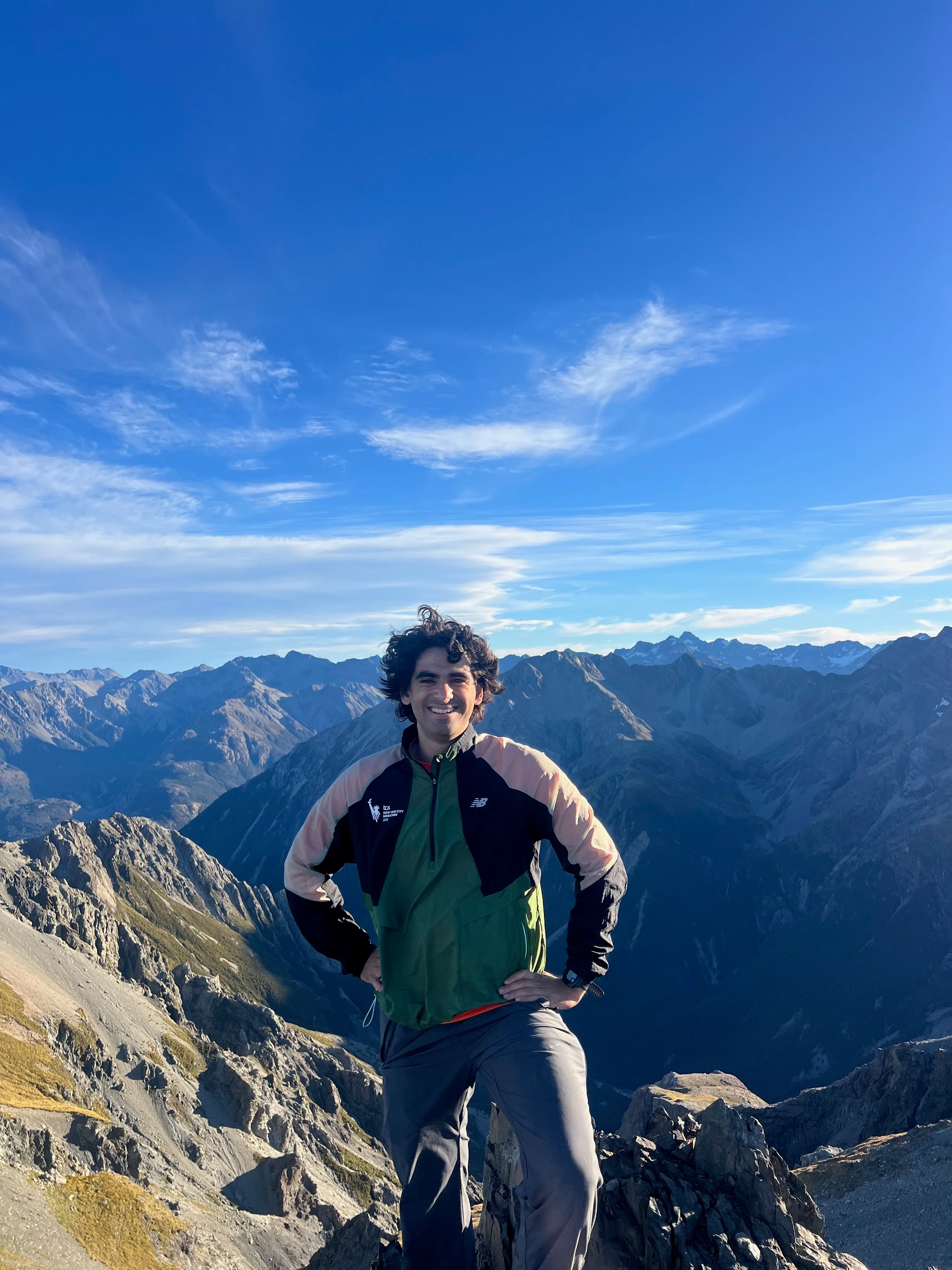 A hiker with hands on hips, smiling on the summit in front of mountain ranges.