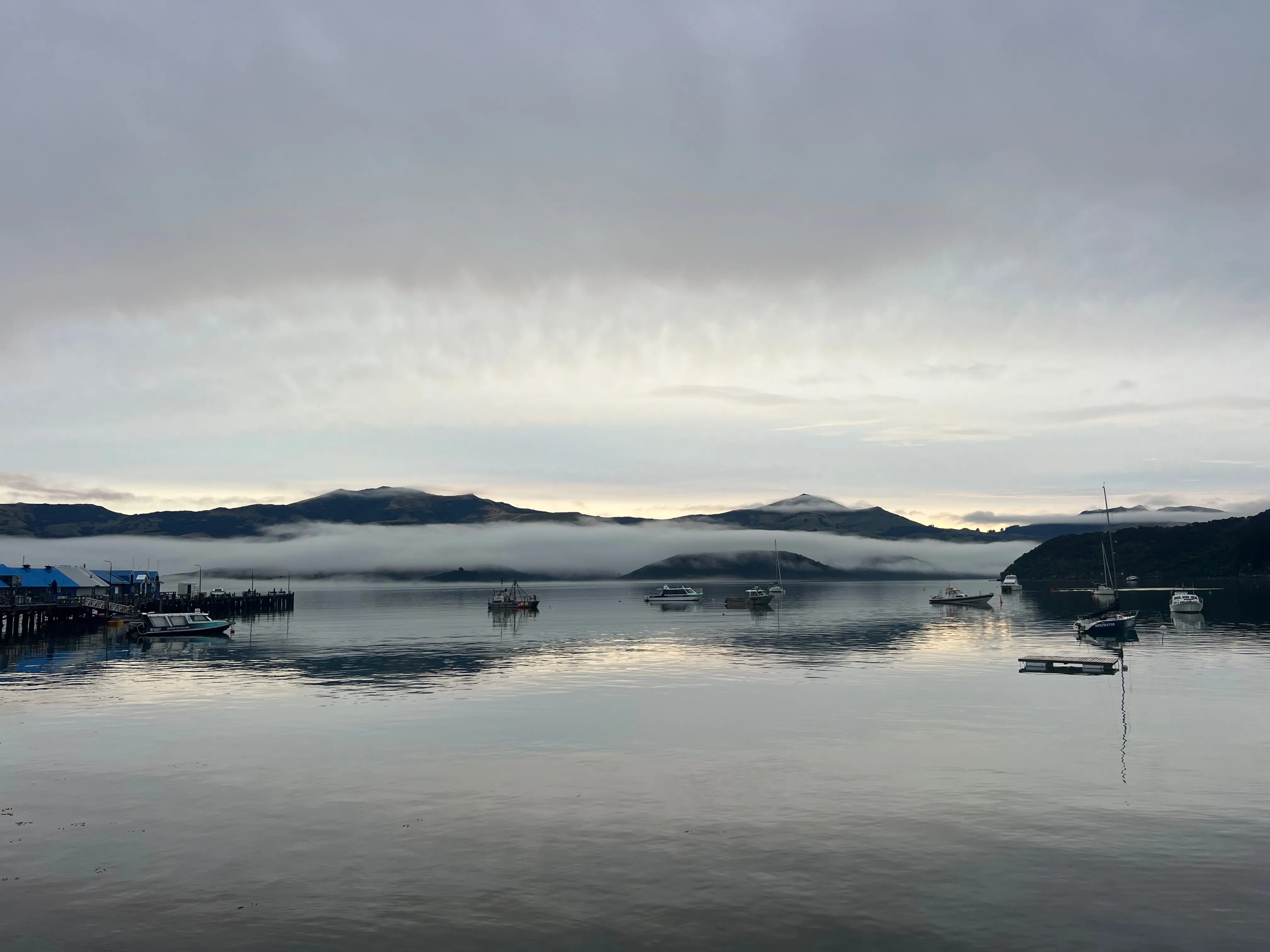 Boats moored on a calm harbour below hills wrapped in low cloud.