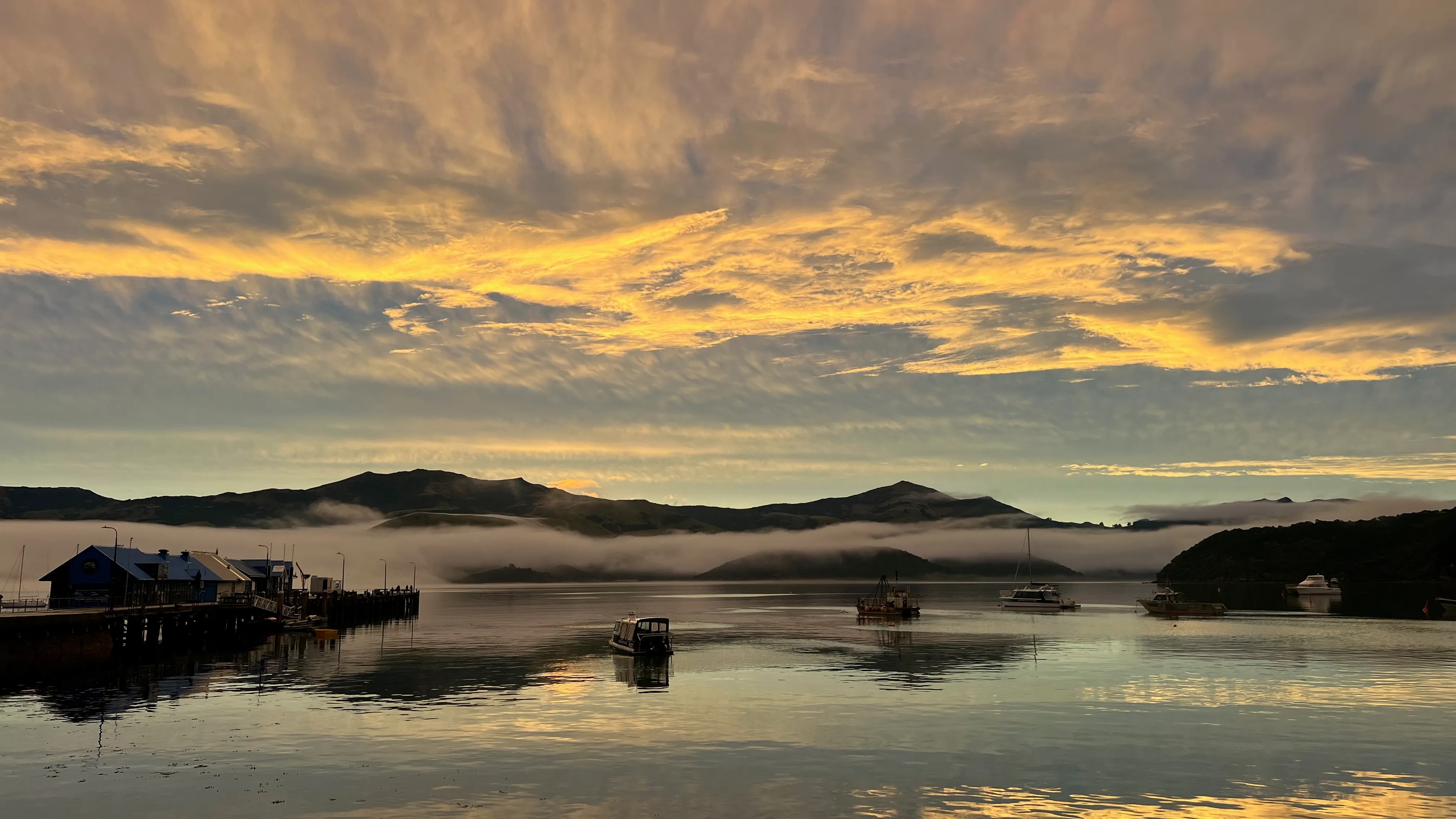A golden evening sky over a wharf and boats on a misty harbour.