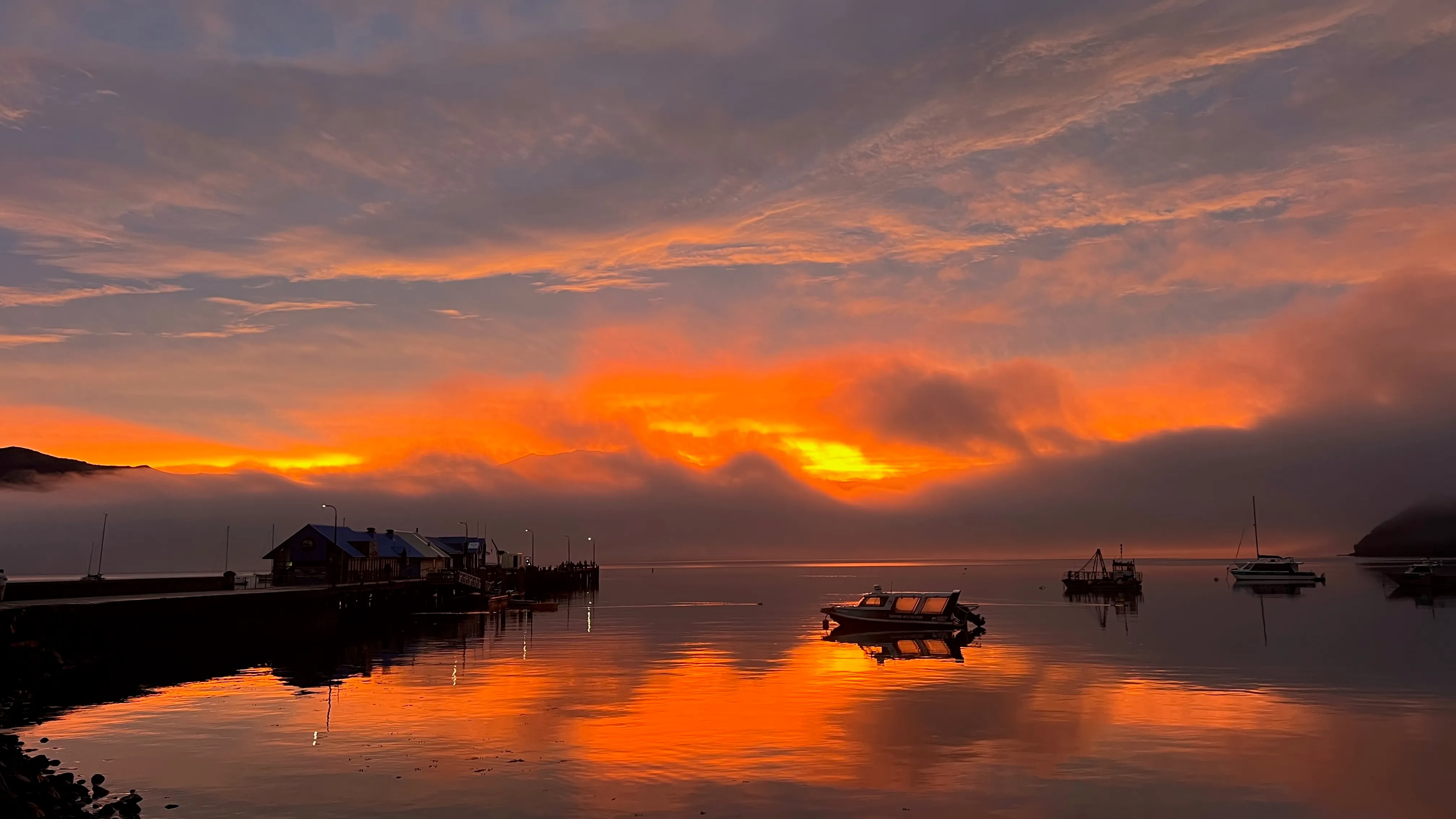 A fiery sunset reflected in the still harbour beside a wharf and moored boats.