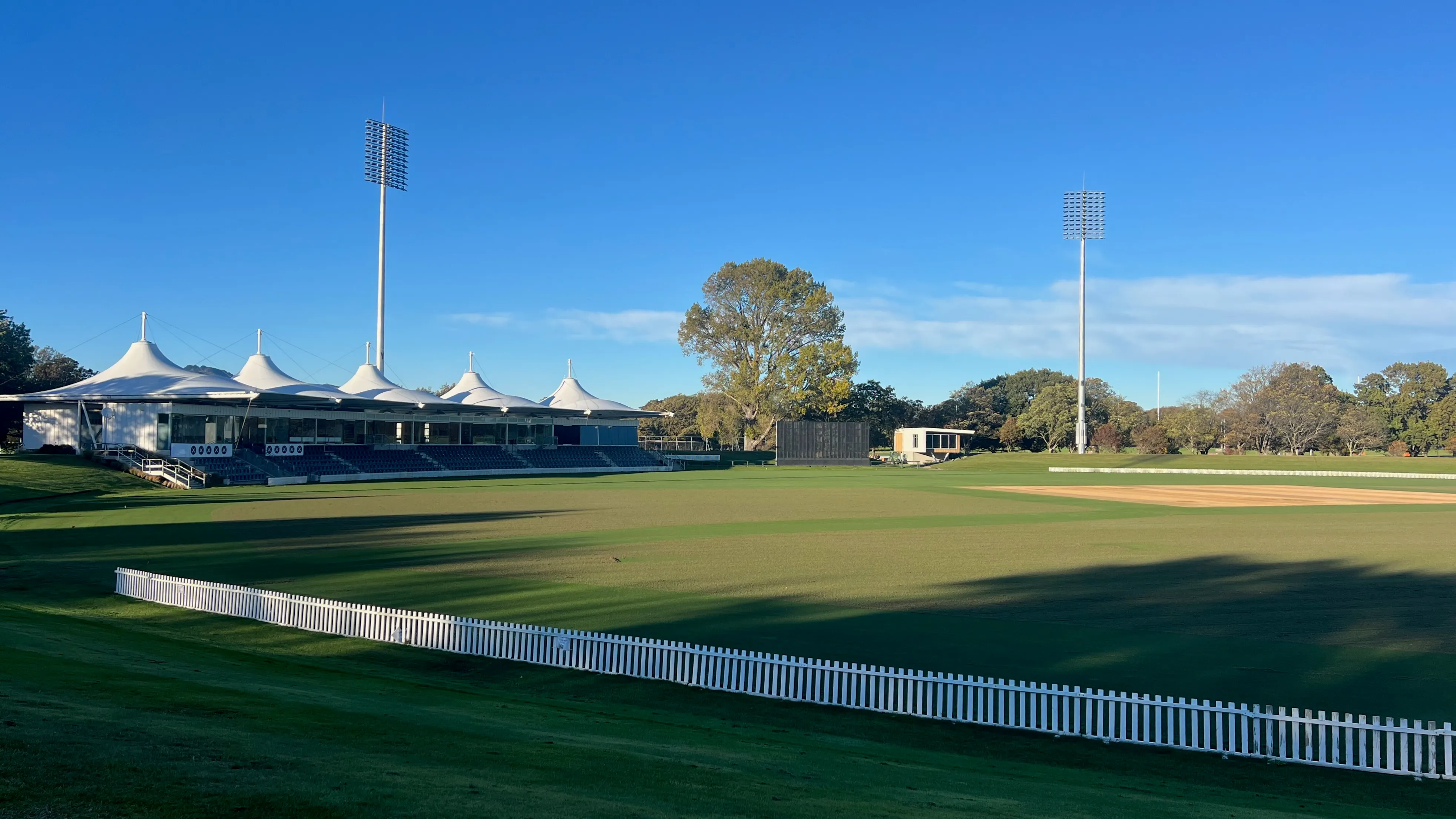 A cricket ground with white sail-roofed stands and a picket fence on a sunny morning.