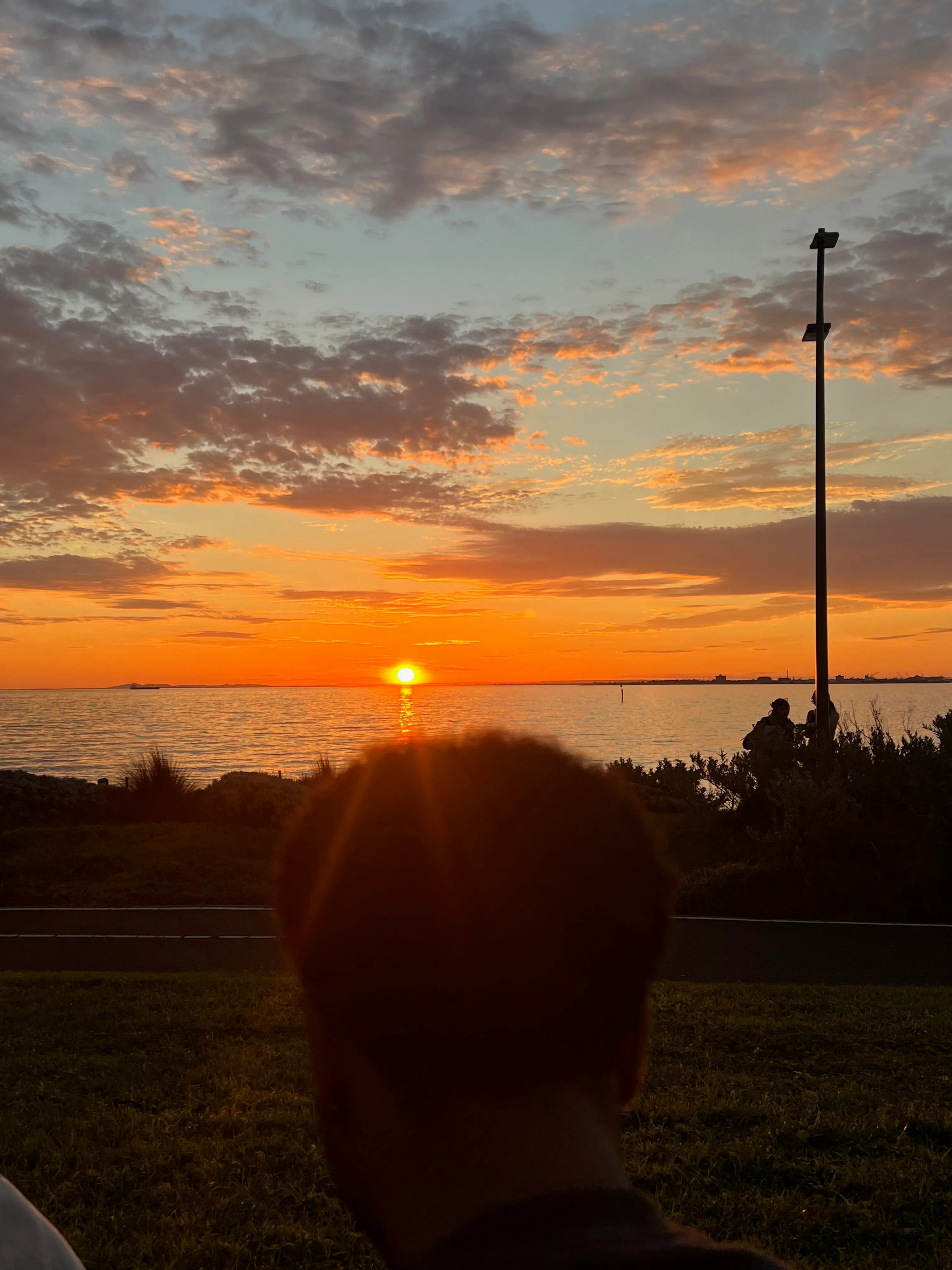 An orange sunset over the bay with people on the grassy foreshore.