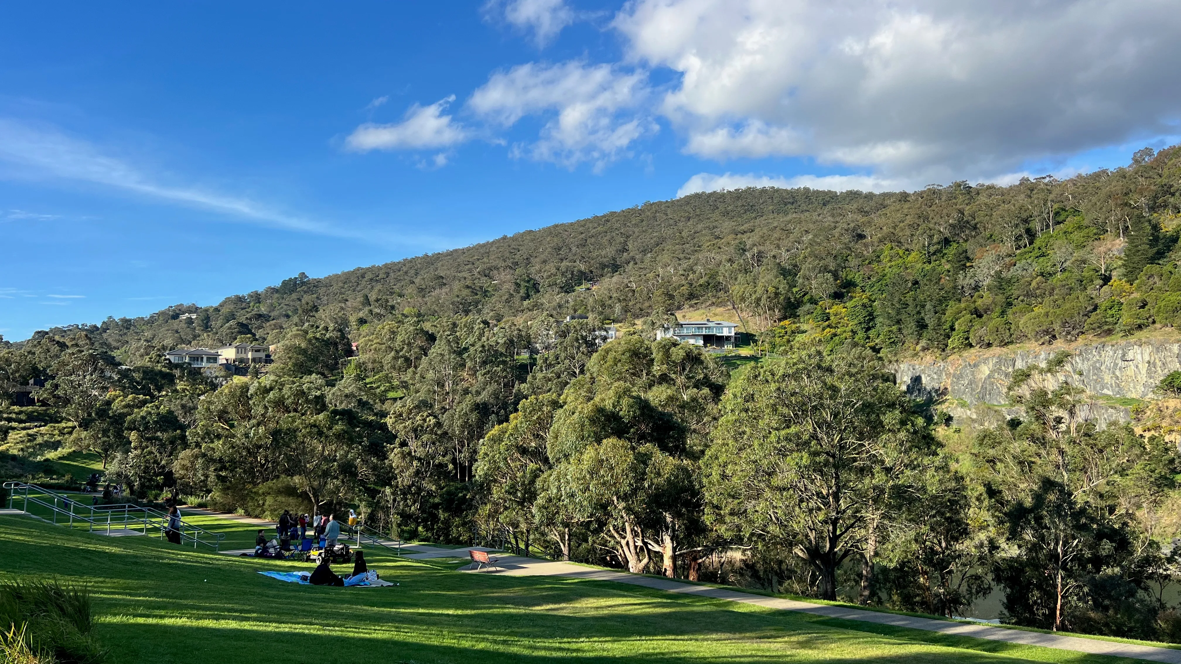 A lakeside park below a wooded hillside on a sunny day.