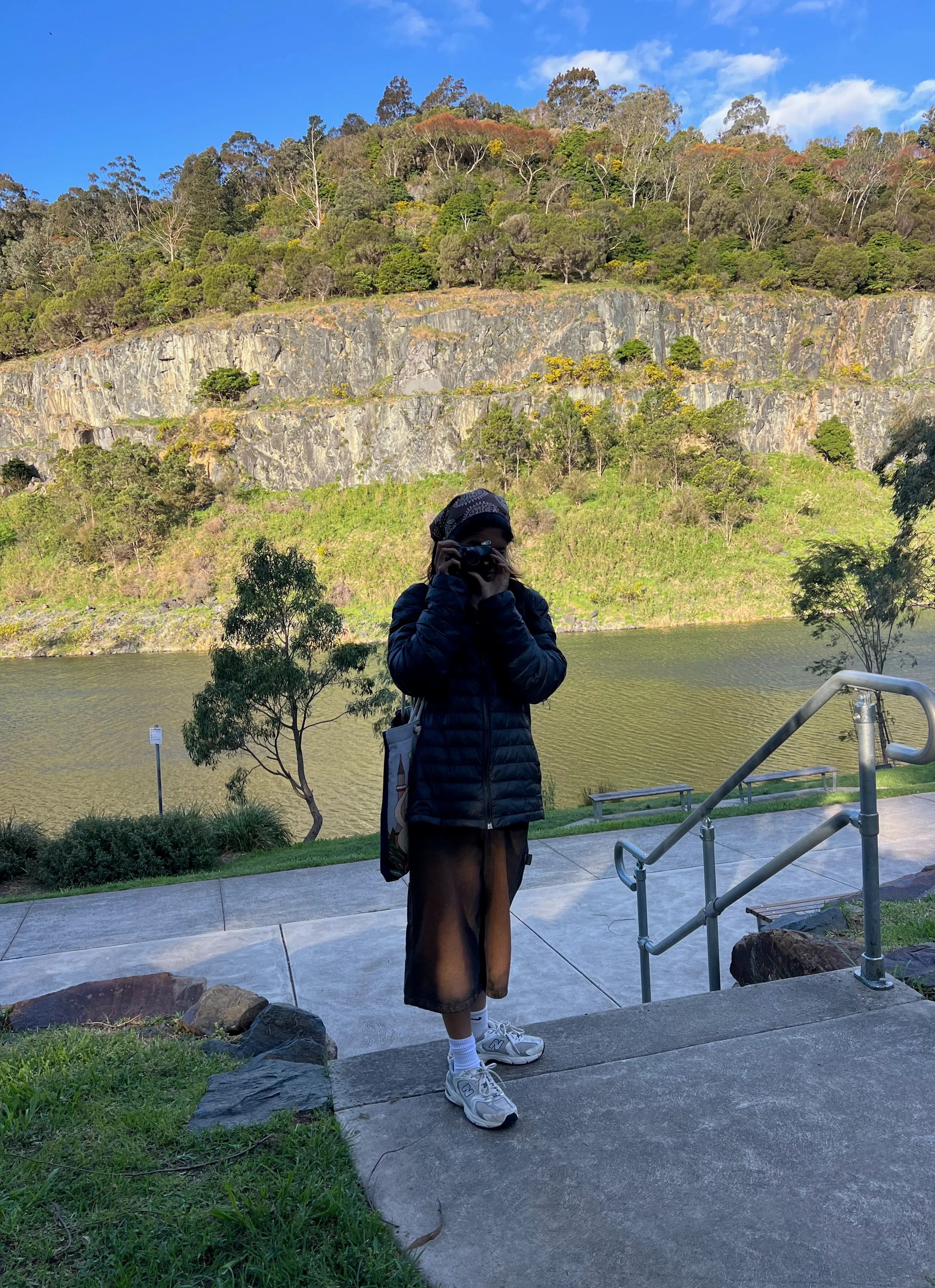 A person holding a camera on steps beside a lake and rocky cliffs.