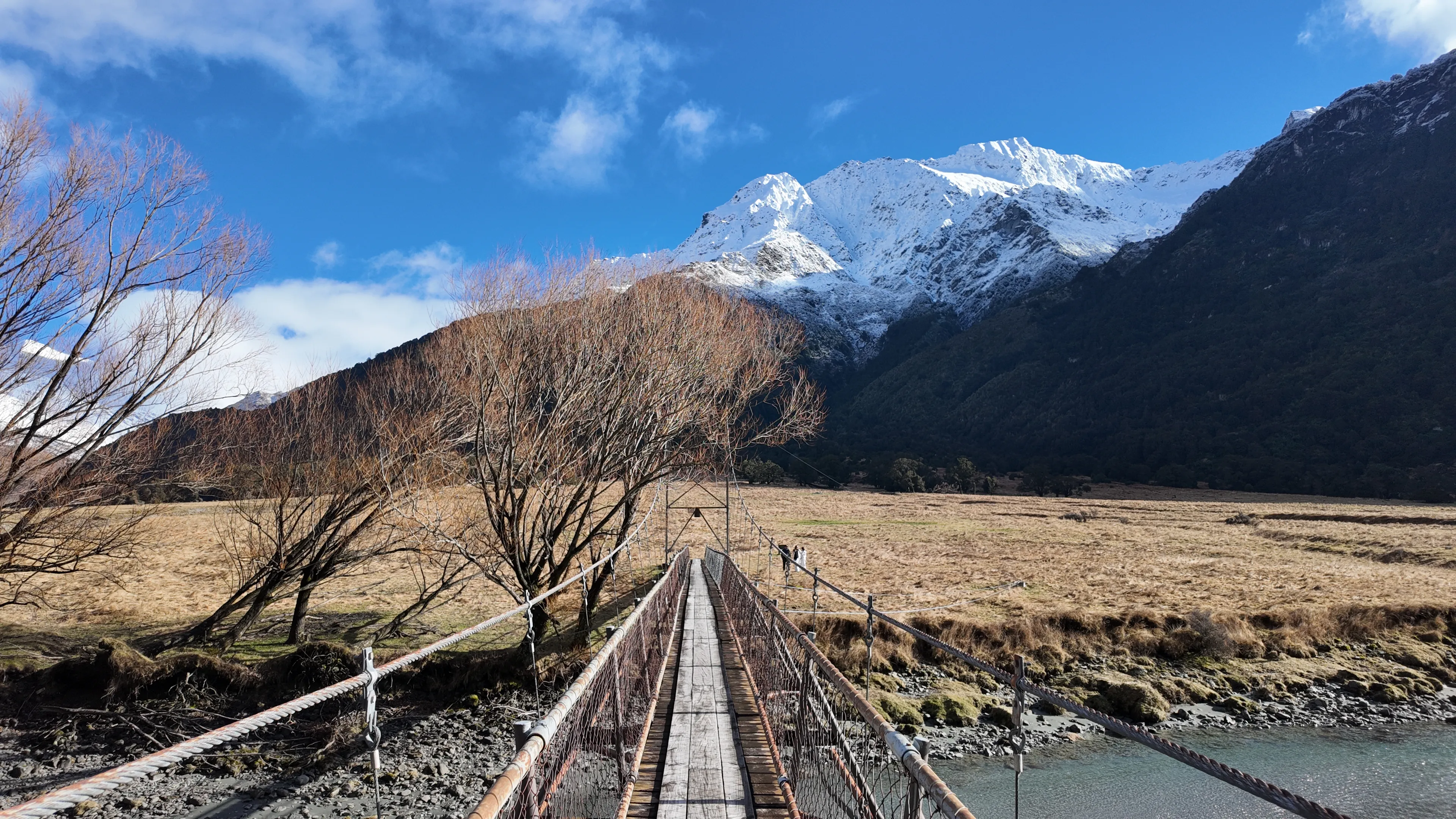 A wooden suspension bridge crossing a river toward a mountain meadow.