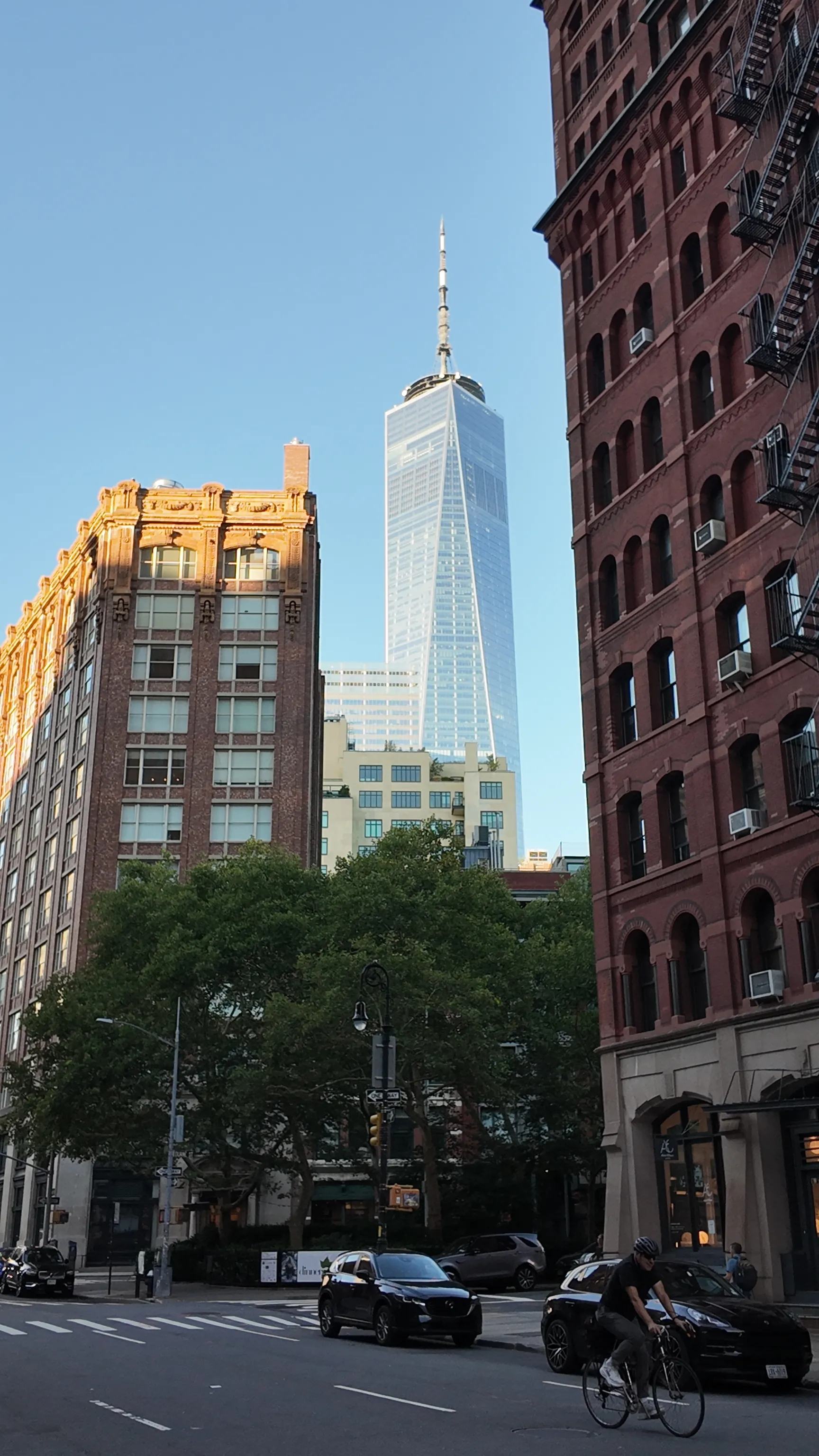 One World Trade Center rising between an ornate brick building and a gilded corner building, with a cyclist crossing the street.