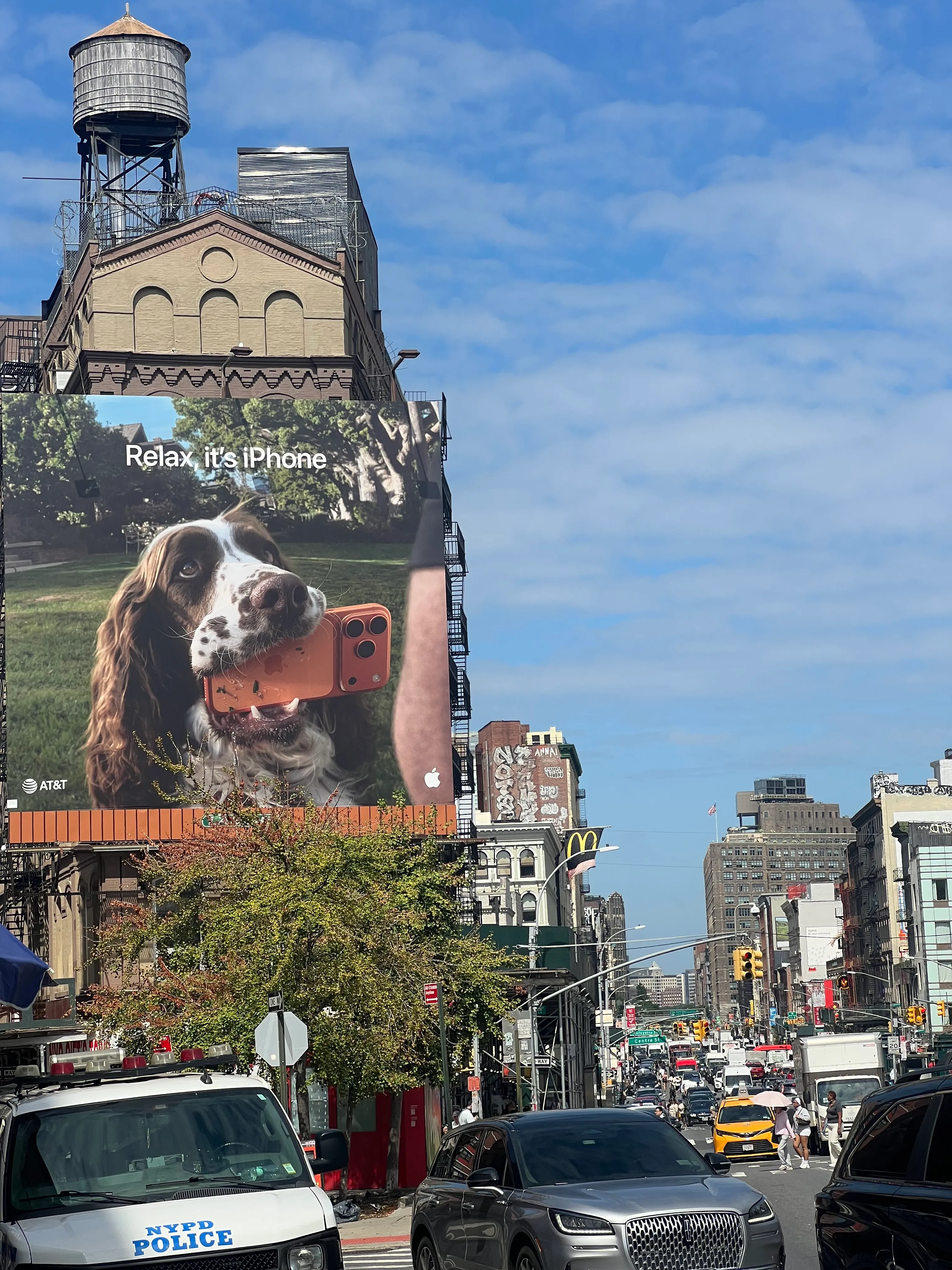 A giant AT&T billboard of a dog holding an iPhone above a busy Centre Street intersection, with an NYPD car in the foreground.