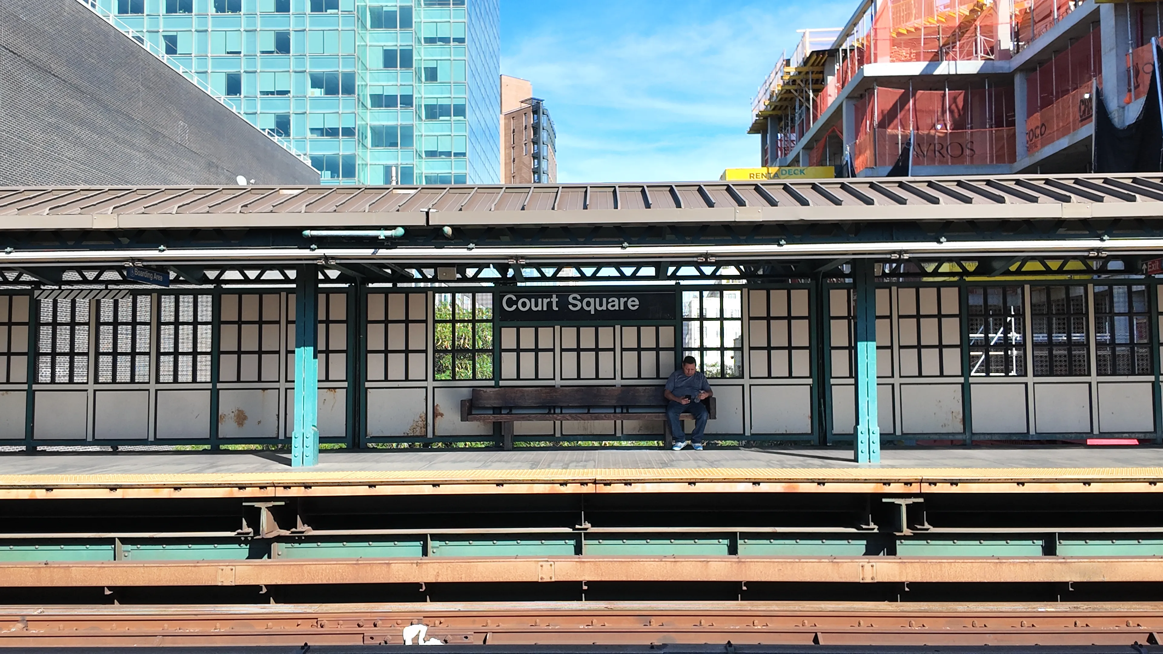 A passenger seated on the Court Square station platform beneath surrounding city buildings.