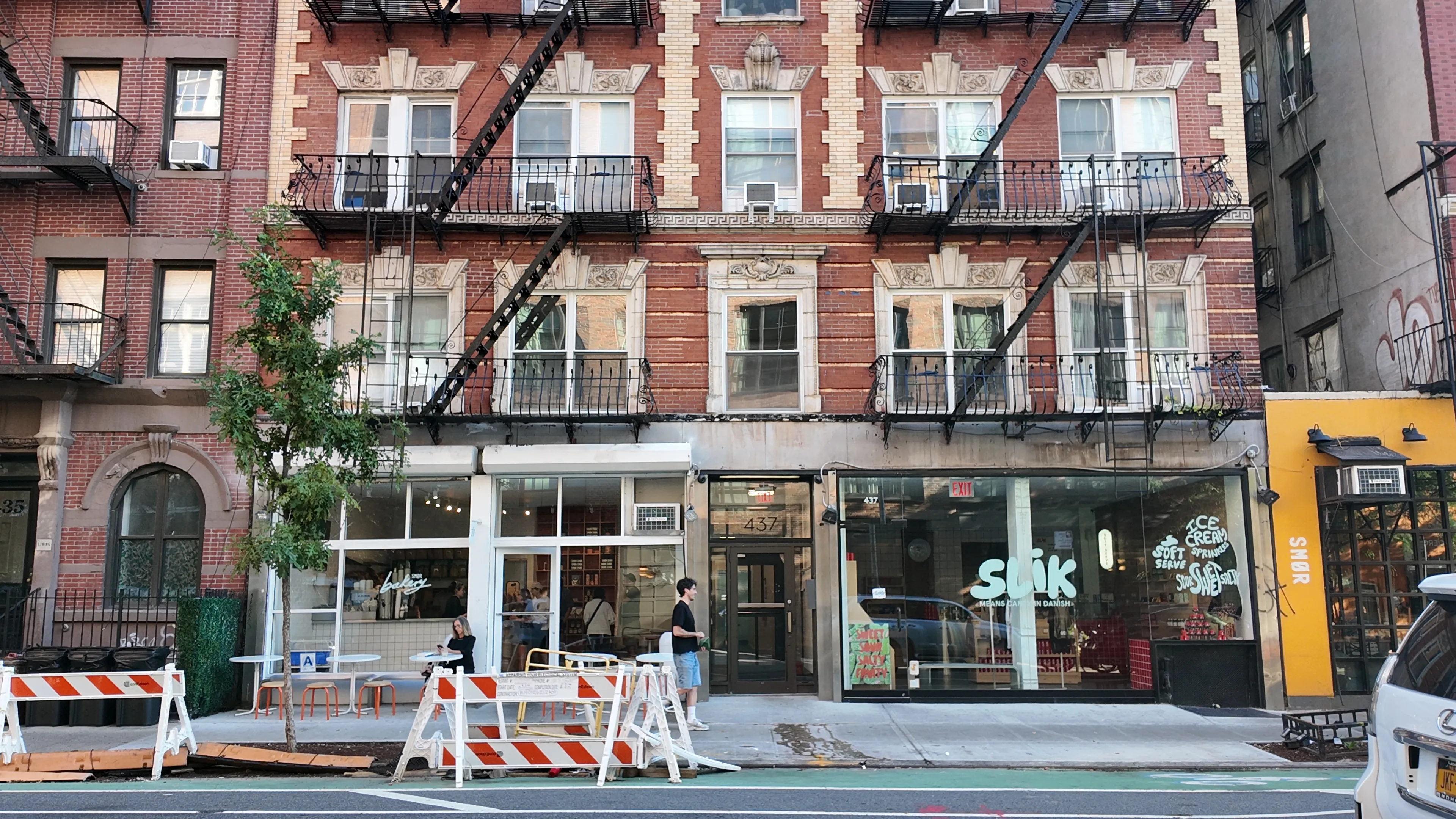 Brick apartment buildings and fire escapes above shopfronts and roadworks.