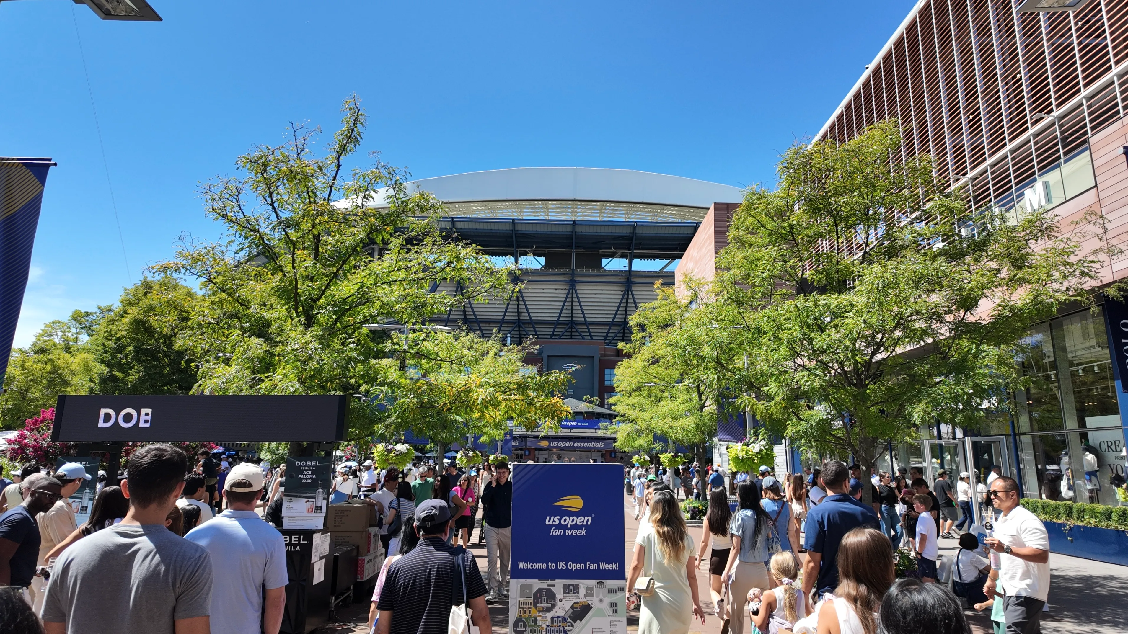 Visitors walking through the US Open grounds beneath trees and stadium buildings.