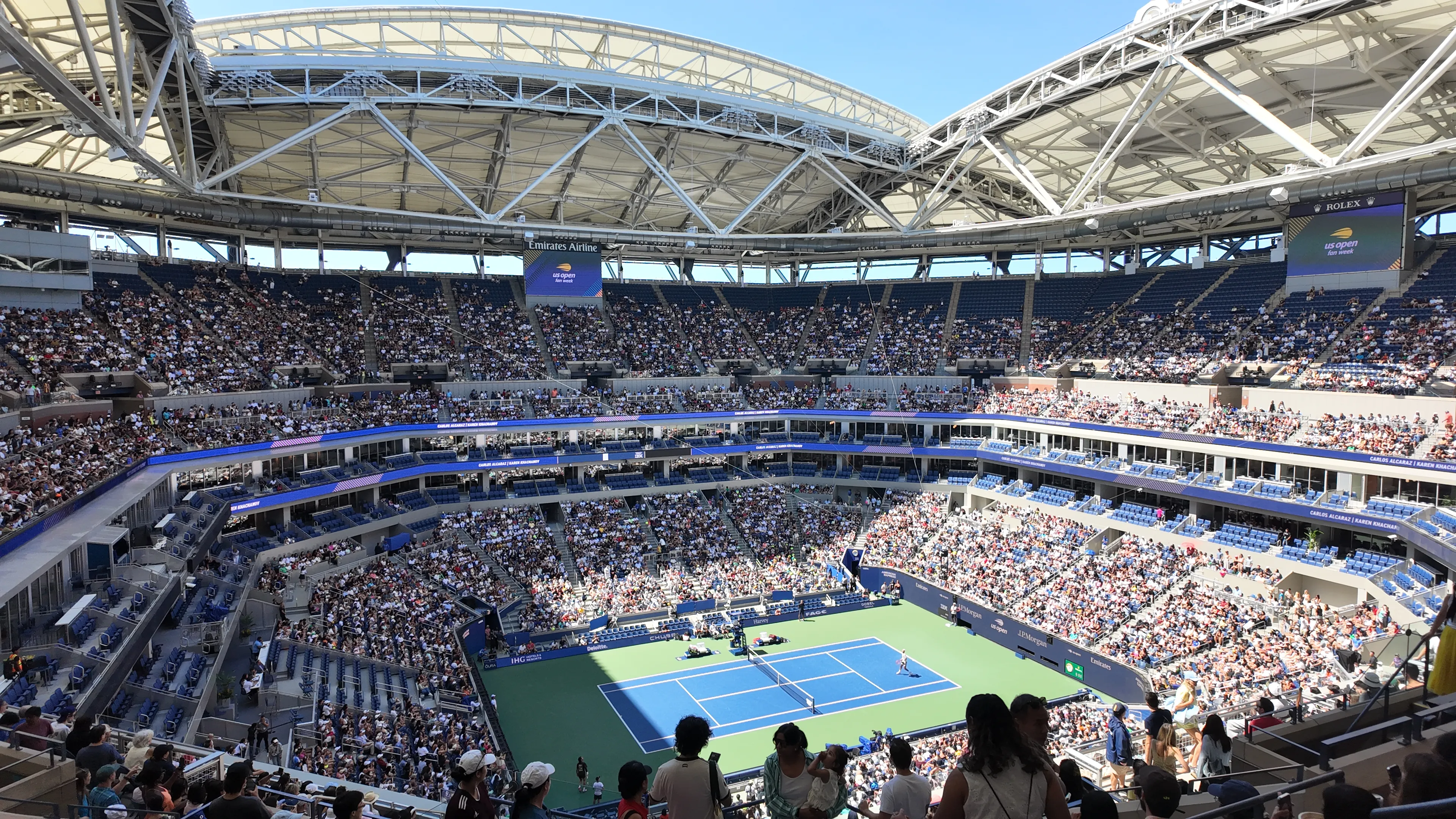A tennis court surrounded by spectators beneath a stadium open roof.