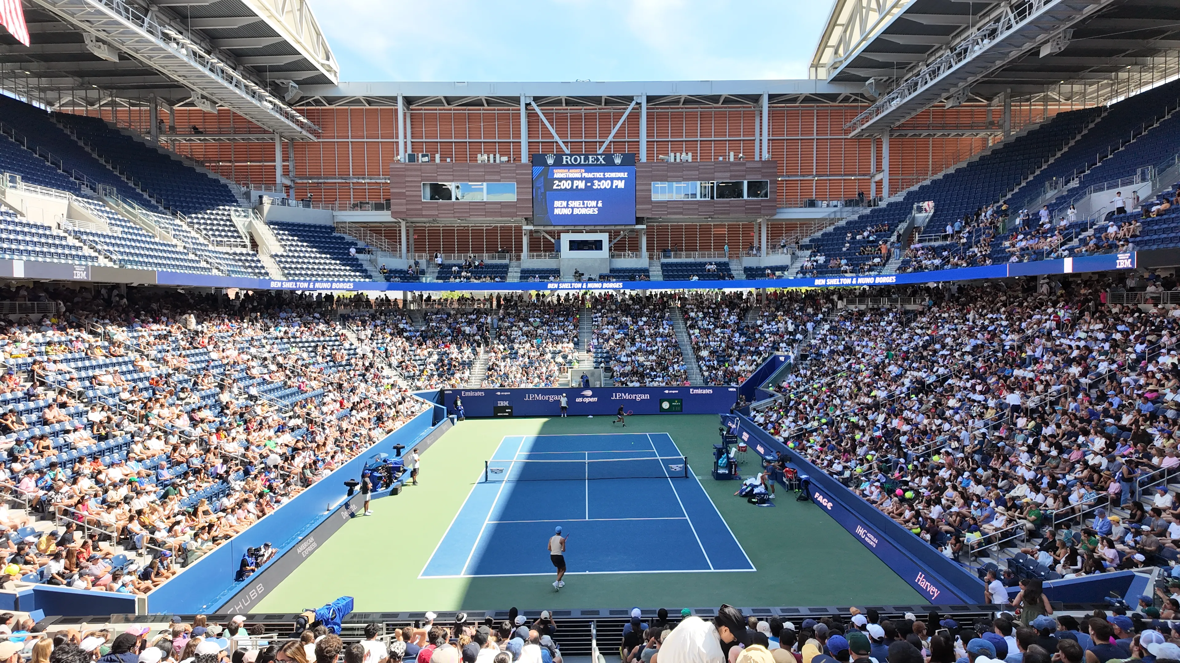Players practising on a blue tennis court with spectators filling the stands.