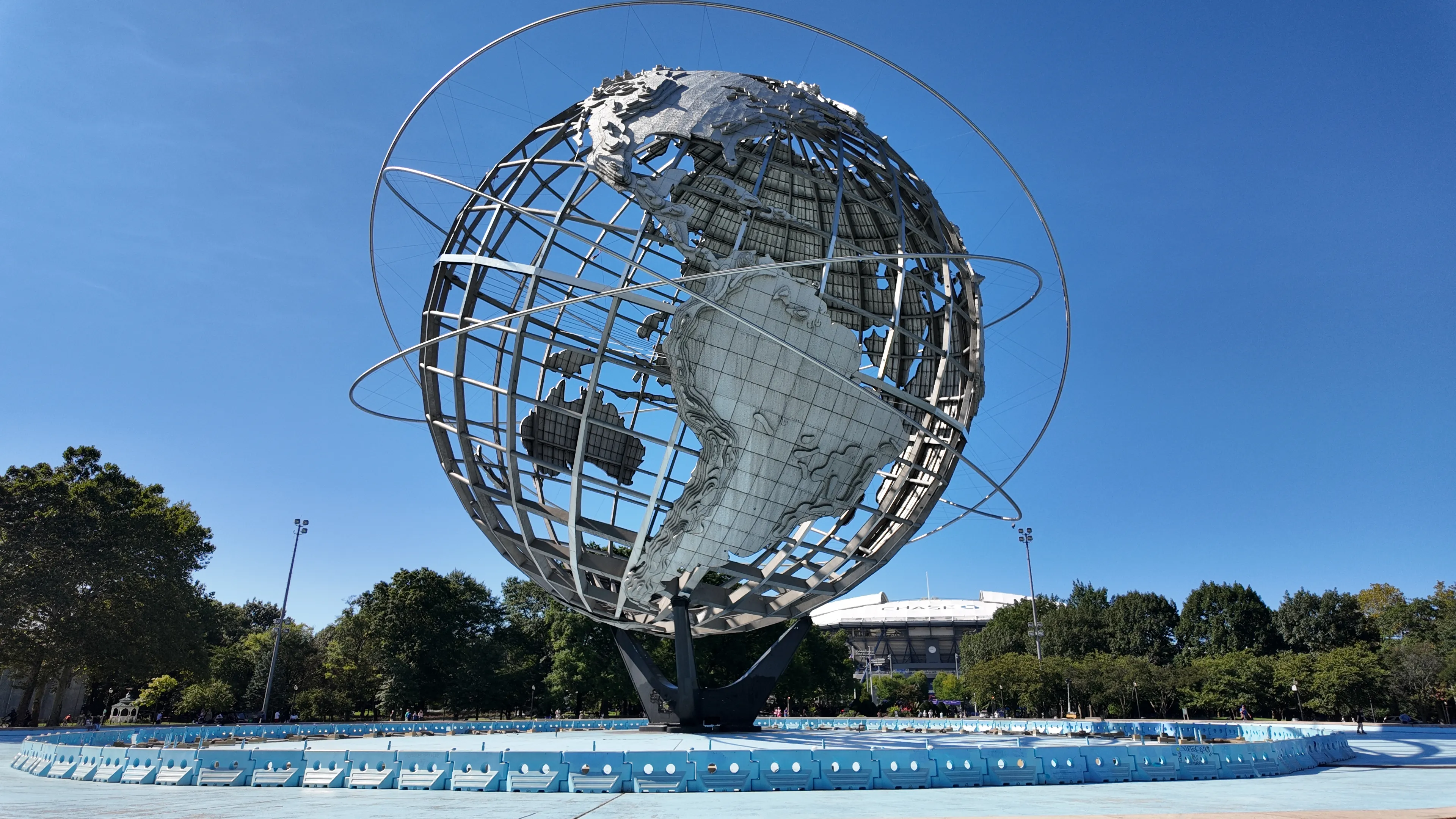 A large steel globe sculpture beside the tennis grounds under a clear blue sky.