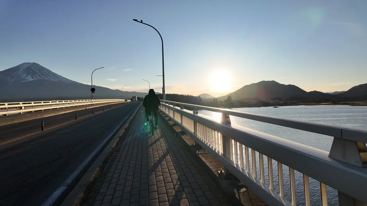 A cyclist on a bridge beside a lake, with a mountain and low sun ahead.