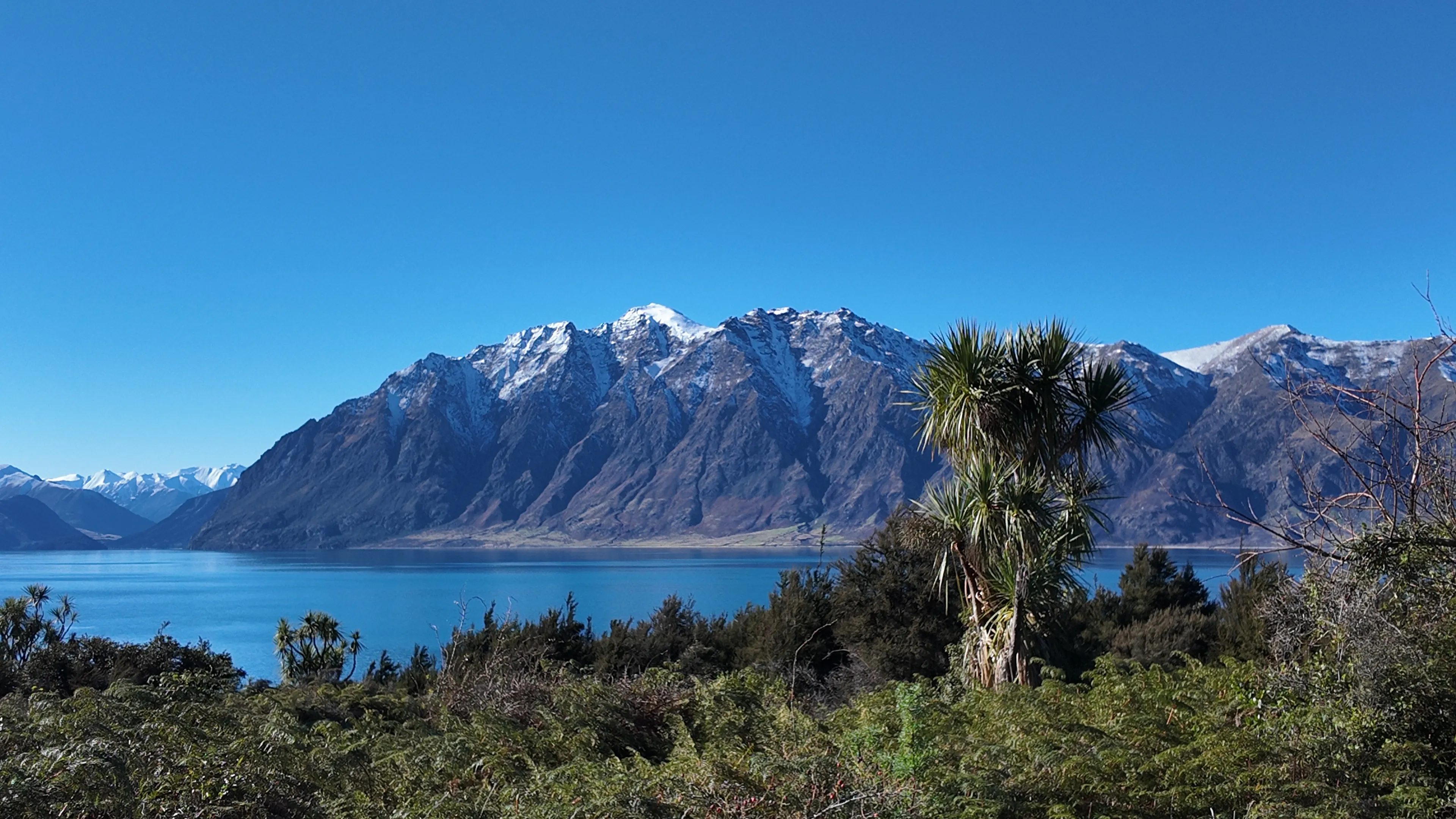 Snow-dusted mountains above a blue lake, with trees in the foreground under a clear sky.