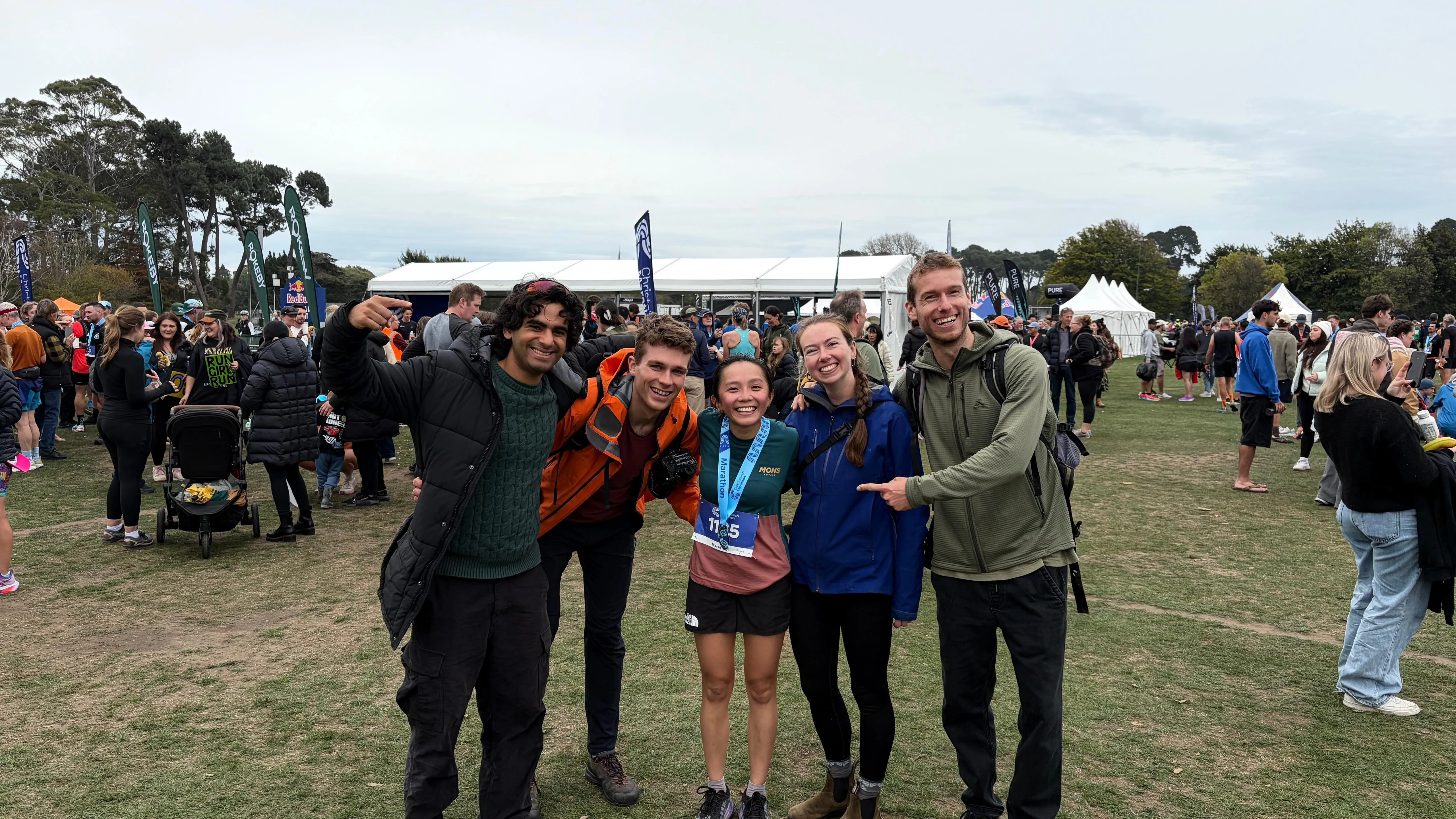 Five friends posing together on a crowded field at a running event, one wearing a finisher's medal.