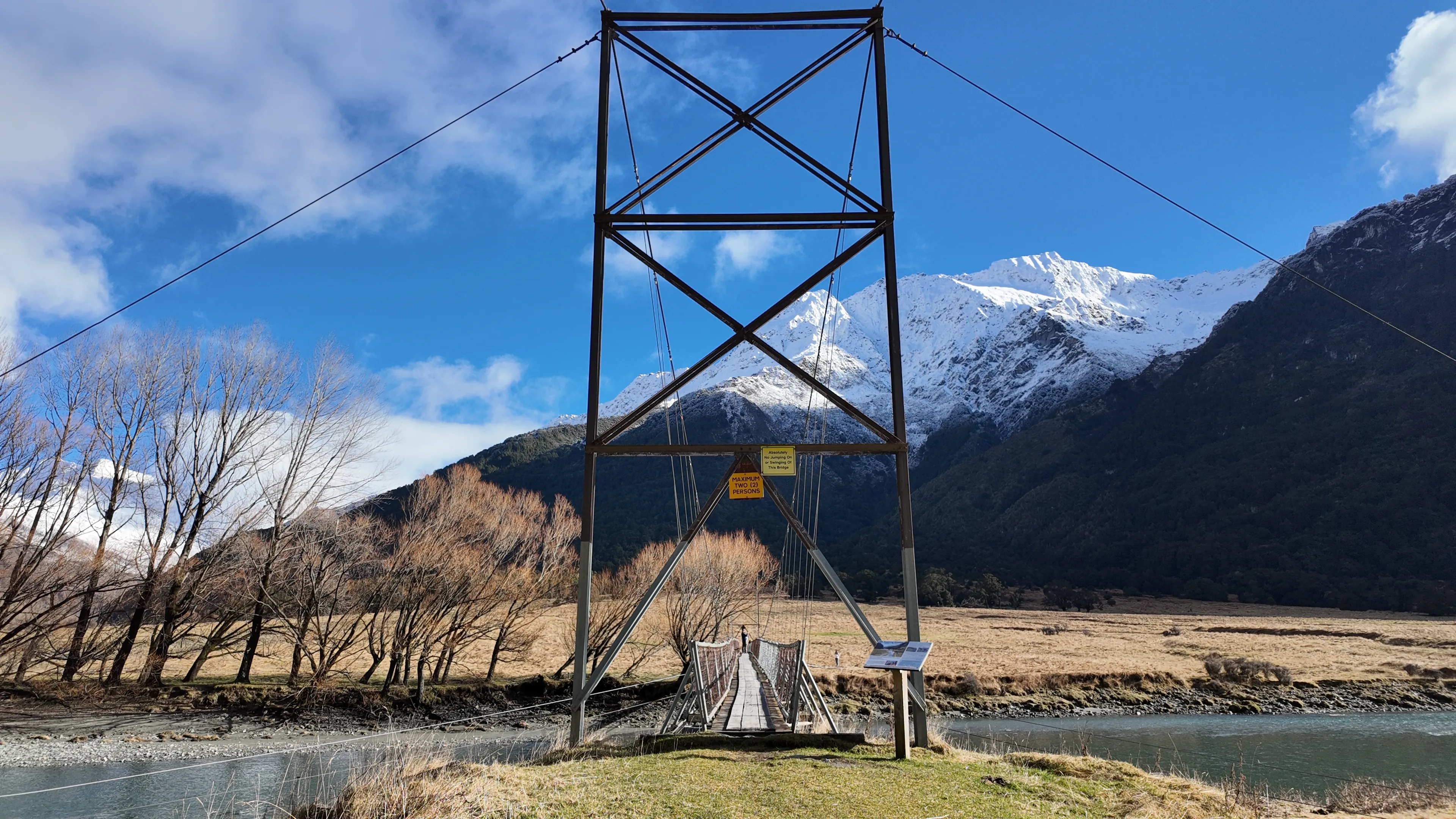 A suspension bridge tower in front of snowy mountains and bare trees.